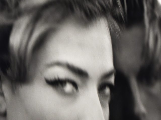 A black and white close-up shot shows a woman's face in the foreground with another person's blurred face behind her.