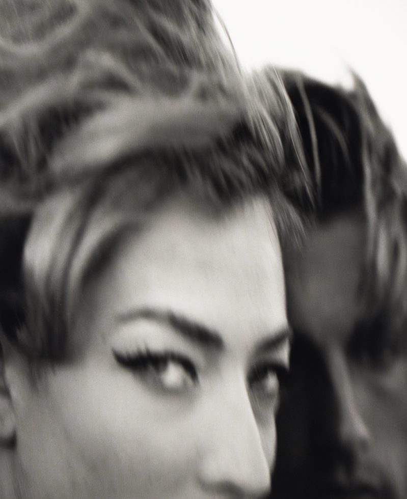 A black and white close-up shot shows a woman's face in the foreground with another person's blurred face behind her.
