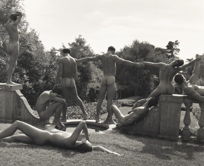 A group of nude figures pose in classical, sculptural stances in a manicured garden with stone architecture and trees in the background.