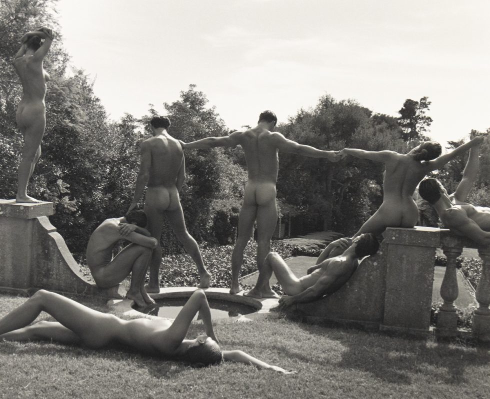 A group of nude figures pose in classical, sculptural stances in a manicured garden with stone architecture and trees in the background.