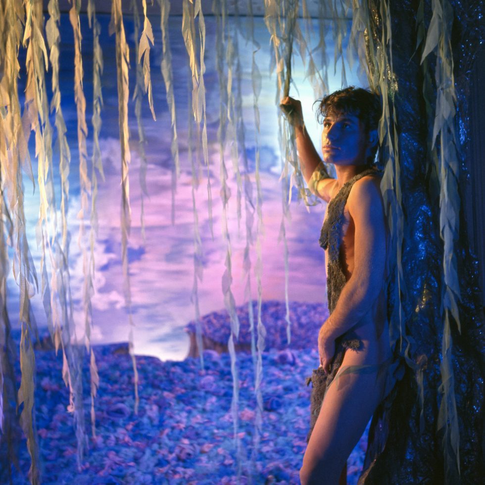 A person leans on a dark tree, partially clad, against a vibrant blue and purple landscape with pink sky and hanging willow-like fabric strips.