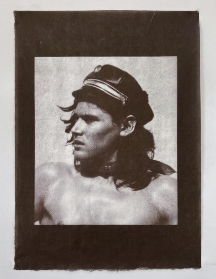 A monochromatic portrait shows a man with long, dark hair wearing a black leather cap. He is shirtless, facing slightly to the left with a serious expression, and the image is framed by a dark border.