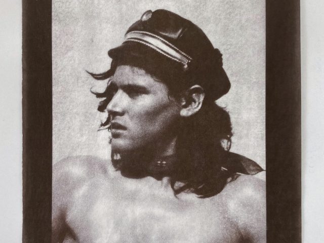 A monochromatic portrait shows a man with long, dark hair wearing a black leather cap. He is shirtless, facing slightly to the left with a serious expression, and the image is framed by a dark border.