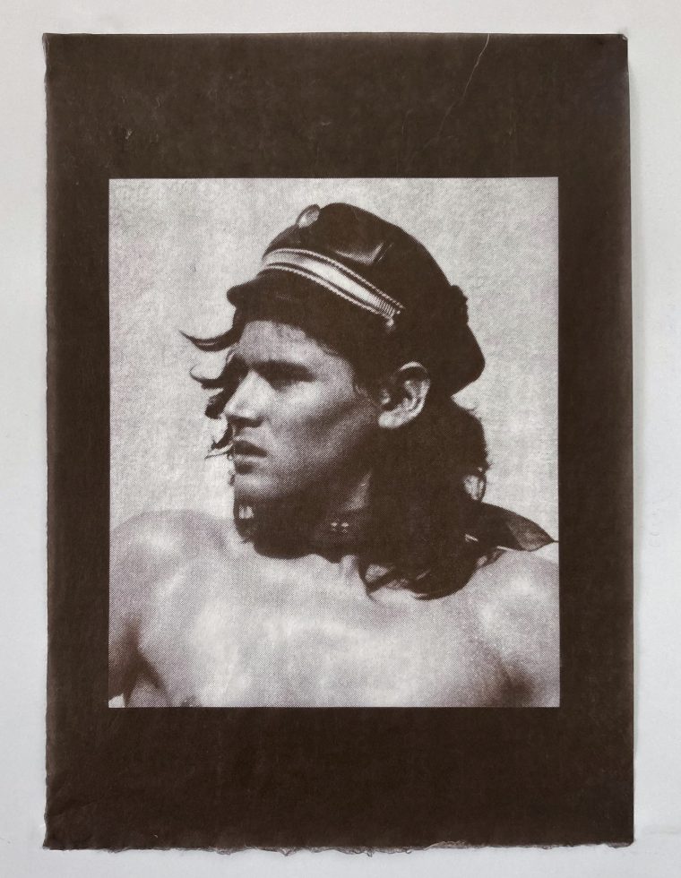 A monochromatic portrait shows a man with long, dark hair wearing a black leather cap. He is shirtless, facing slightly to the left with a serious expression, and the image is framed by a dark border.