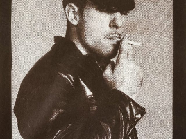 A male figure in a leather outfit smoking a cigarette.