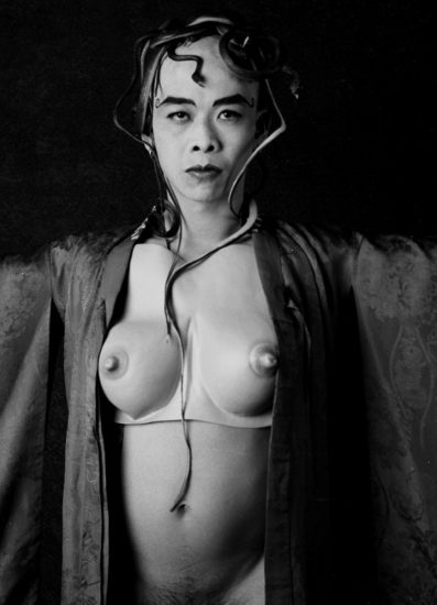 A person with a serious expression and snakes woven into their hair and draped over their body, wearing a robe over a chest plate with prominent nipples.