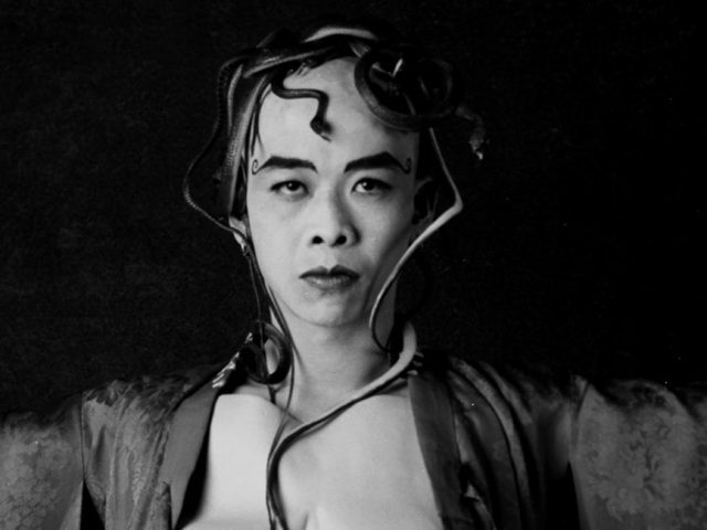 A person with a serious expression and snakes woven into their hair and draped over their body, wearing a robe over a chest plate with prominent nipples.