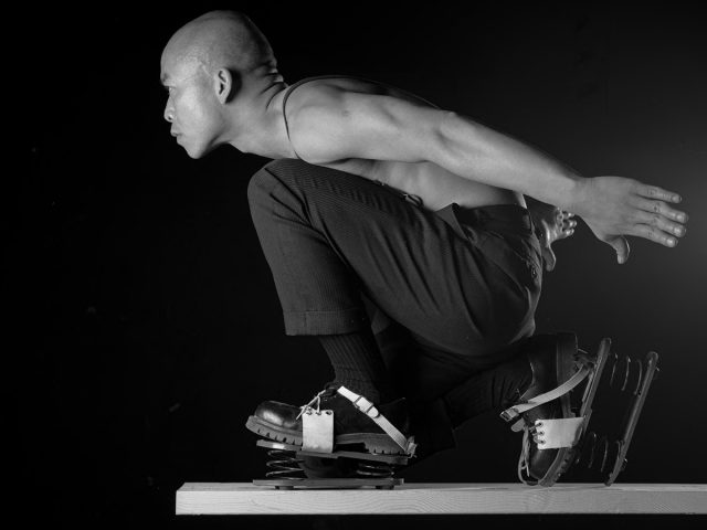 A man in a crouched position, wearing spring-loaded shoes, is balanced on a wooden plank against a black background.