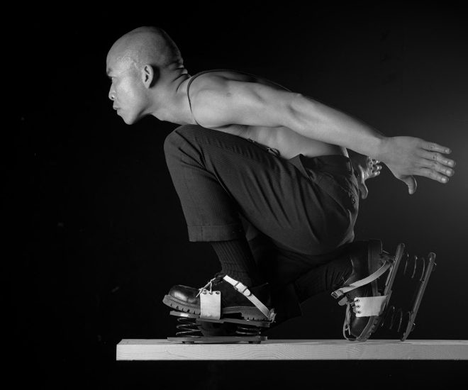 A man in a crouched position, wearing spring-loaded shoes, is balanced on a wooden plank against a black background.