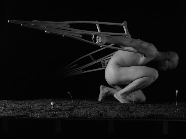 A bald, nude person crouches, with a pair of crutches attached to their back like wings, against a dark background with small plants growing.
