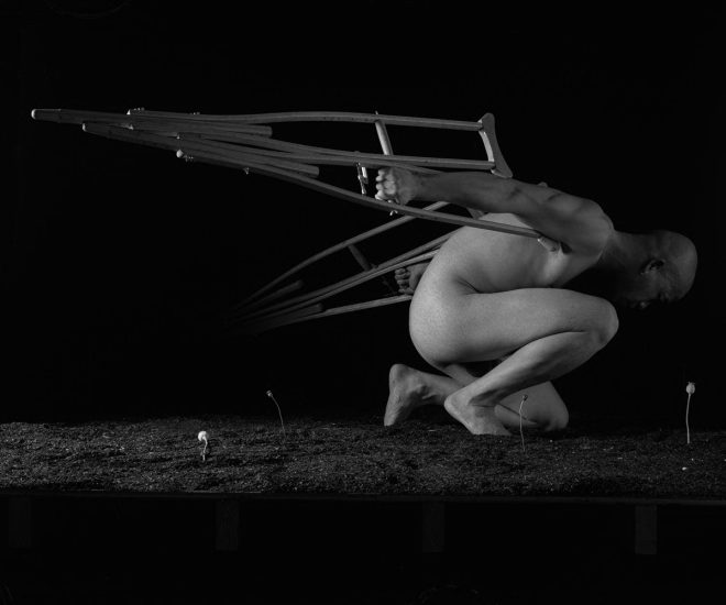 A bald, nude person crouches, with a pair of crutches attached to their back like wings, against a dark background with small plants growing.