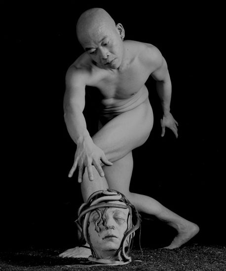 A black-and-white, surreal image of a bald, nude man in a twisted pose, reaching toward a Medusa-like severed head with snakes on the ground.