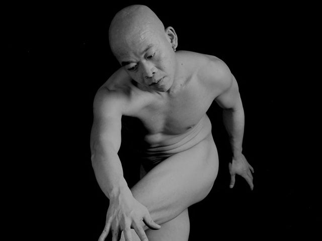 A black-and-white, surreal image of a bald, nude man in a twisted pose, reaching toward a Medusa-like severed head with snakes on the ground.