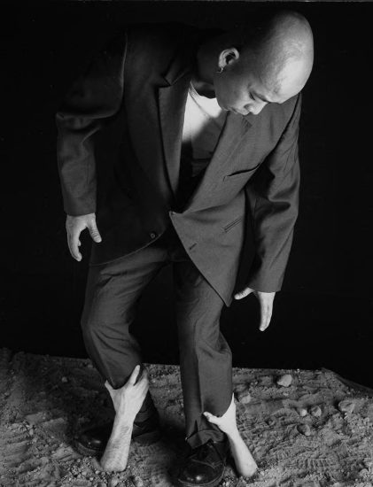 A man in a suit looks down as two disembodied hands grip his ankles, emerging from a surreal, textured, sandy ground.
