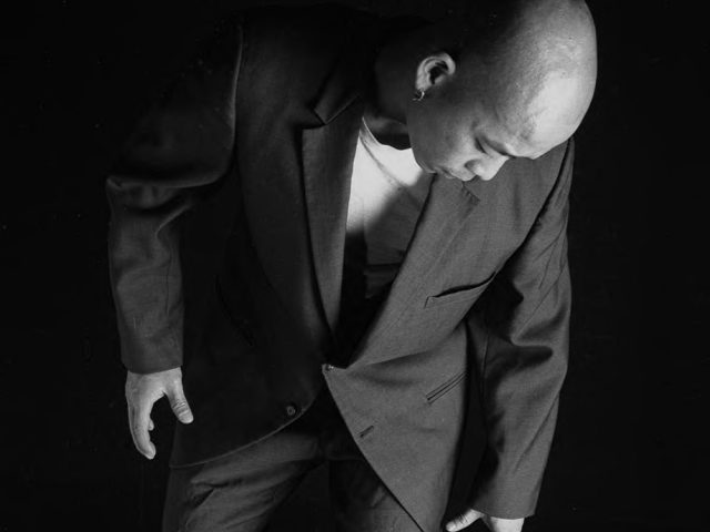 A man in a suit looks down as two disembodied hands grip his ankles, emerging from a surreal, textured, sandy ground.