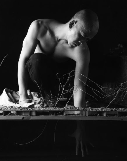 A surreal black and white image of a bald man crouching over a small plot of dirt and tangled branches, with his reflection visible underneath.