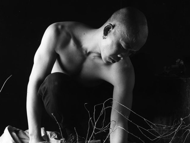 A surreal black and white image of a bald man crouching over a small plot of dirt and tangled branches, with his reflection visible underneath.