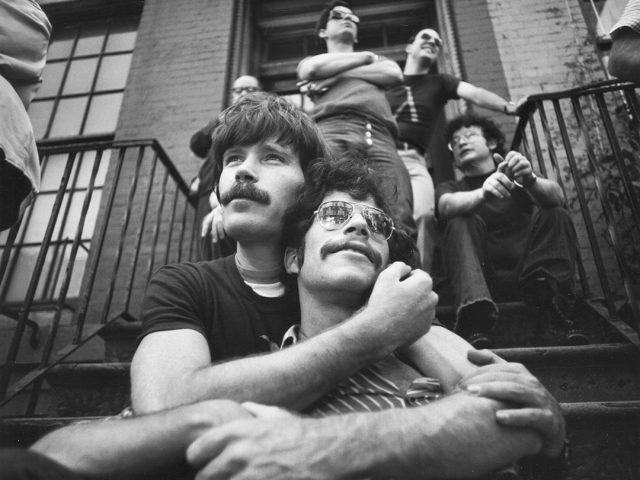 A black and white photograph of two men with mustaches embracing on the steps of a brownstone.