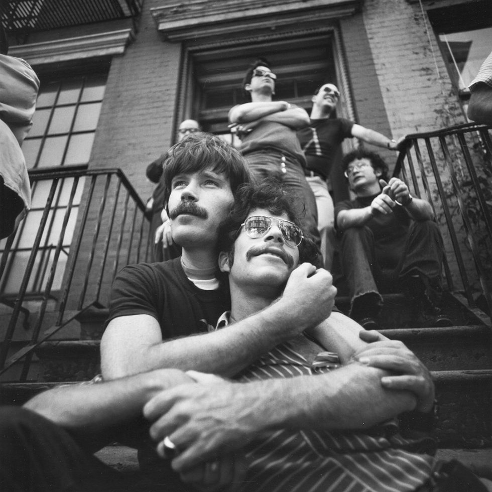 A black and white photograph of two men with mustaches embracing on the steps of a brownstone.