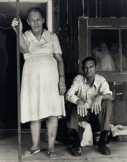 An elderly woman stands on a wooden porch beside a seated man, while a man and child are visible through a nearby window.