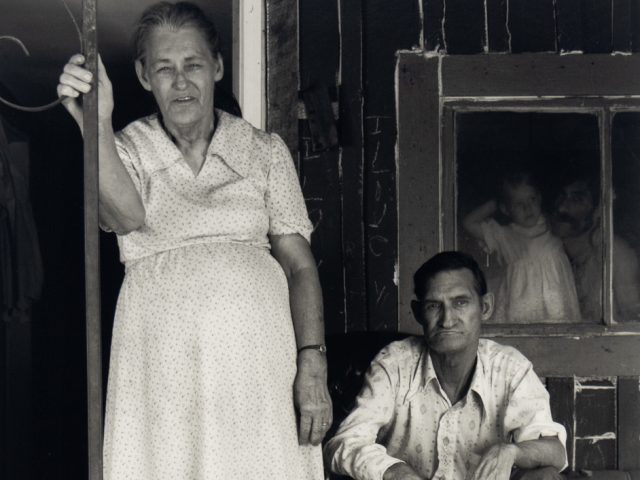An elderly woman stands on a wooden porch beside a seated man, while a man and child are visible through a nearby window.