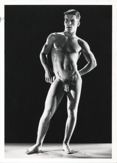 A black-and-white photograph shows a nude man, standing and posing against a dark background, with his hands on his hips.