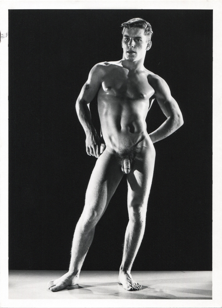 A black-and-white photograph shows a nude man, standing and posing against a dark background, with his hands on his hips.