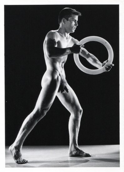 Nude figure stands in a wide pose holding a circular object, muscles highlighted by dramatic lighting against a dark background.