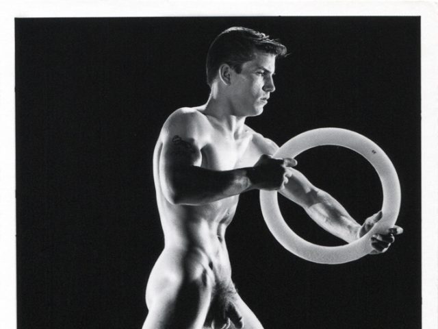 Nude figure stands in a wide pose holding a circular object, muscles highlighted by dramatic lighting against a dark background.
