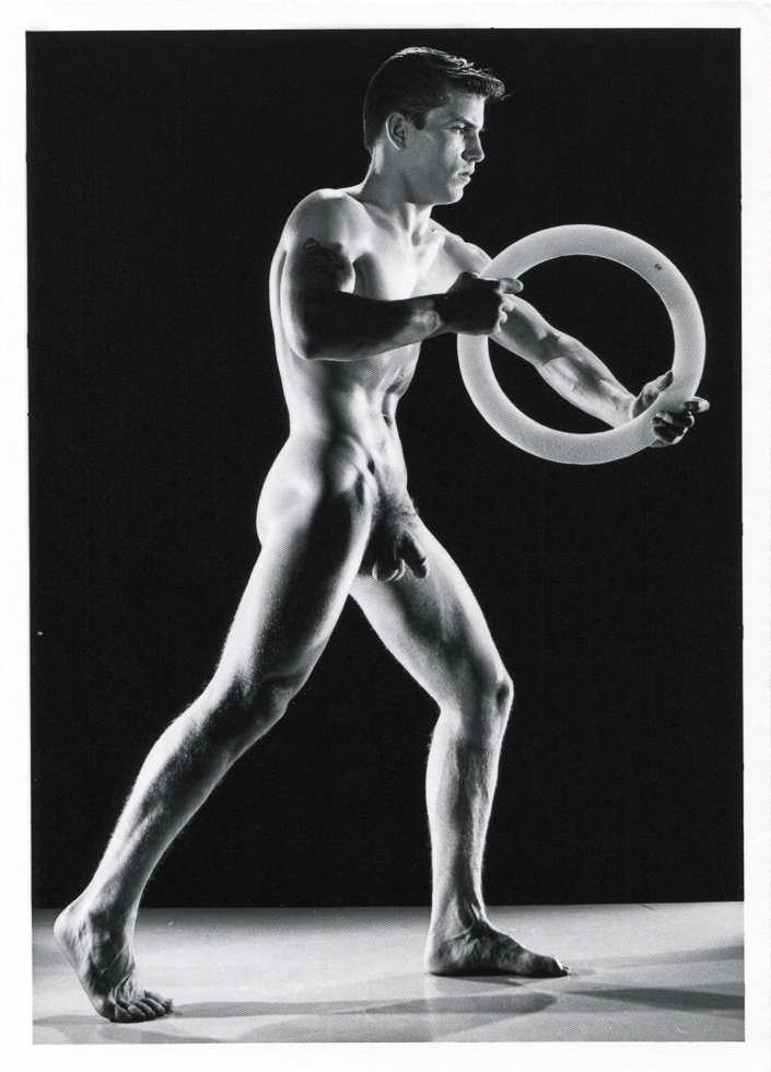 Nude figure stands in a wide pose holding a circular object, muscles highlighted by dramatic lighting against a dark background.