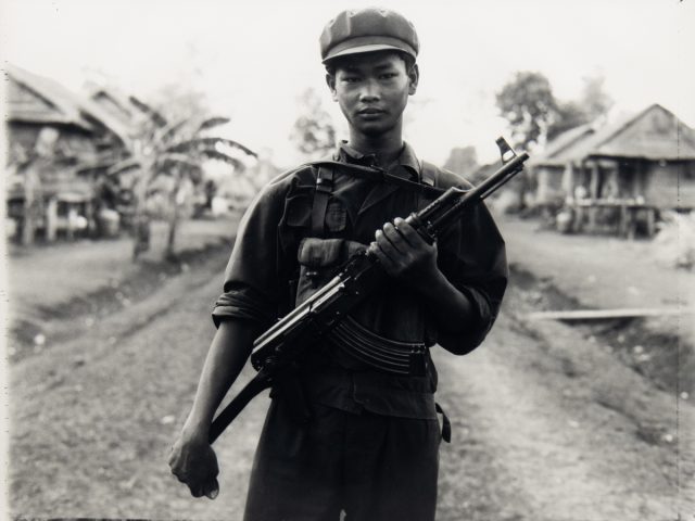 A young soldier in uniform stands on a dirt path in a rural village, holding a rifle across his chest with houses and trees in the background