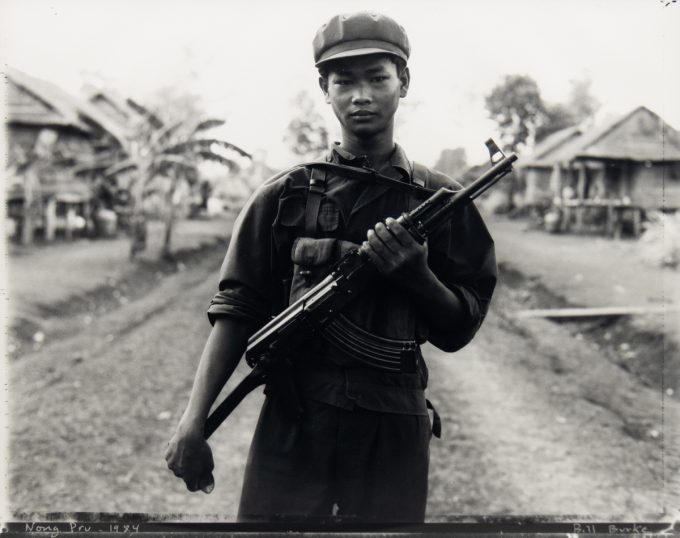 A young soldier in uniform stands on a dirt path in a rural village, holding a rifle across his chest with houses and trees in the background