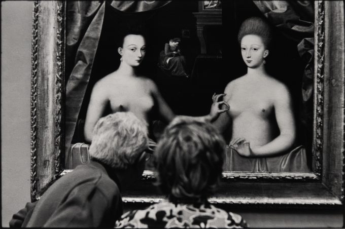 Two people view a painting of two women, creating a playful overlap between the gallery scene and the artwork.