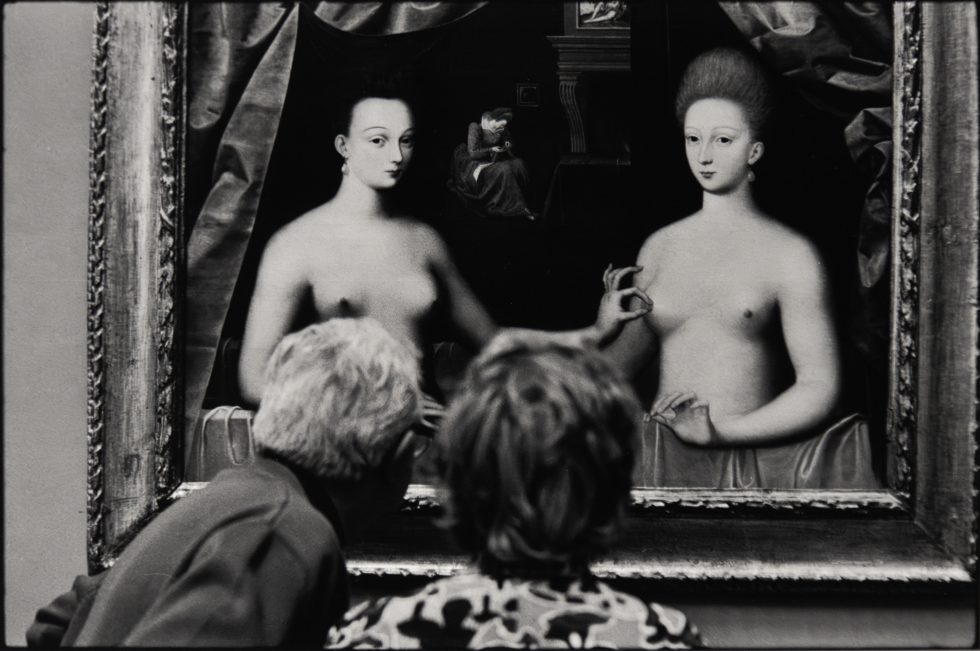 Two people view a painting of two women, creating a playful overlap between the gallery scene and the artwork.
