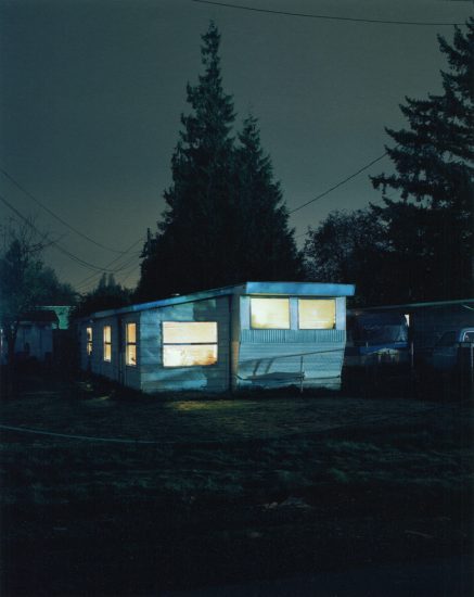 A trailer at night glows with interior light, its pale siding and windows stark against surrounding trees and deep shadows.