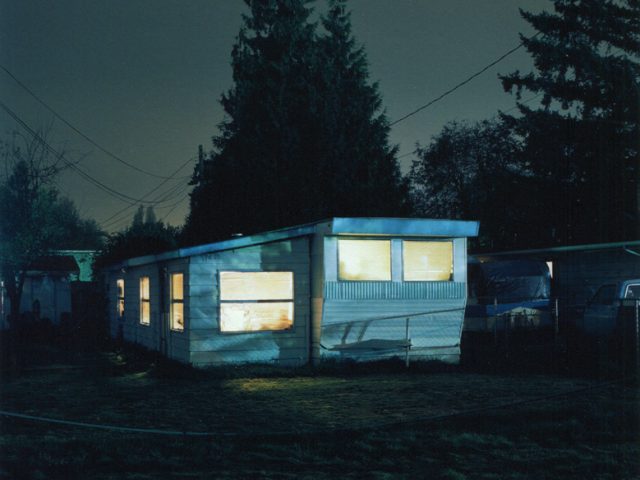 A trailer at night glows with interior light, its pale siding and windows stark against surrounding trees and deep shadows.