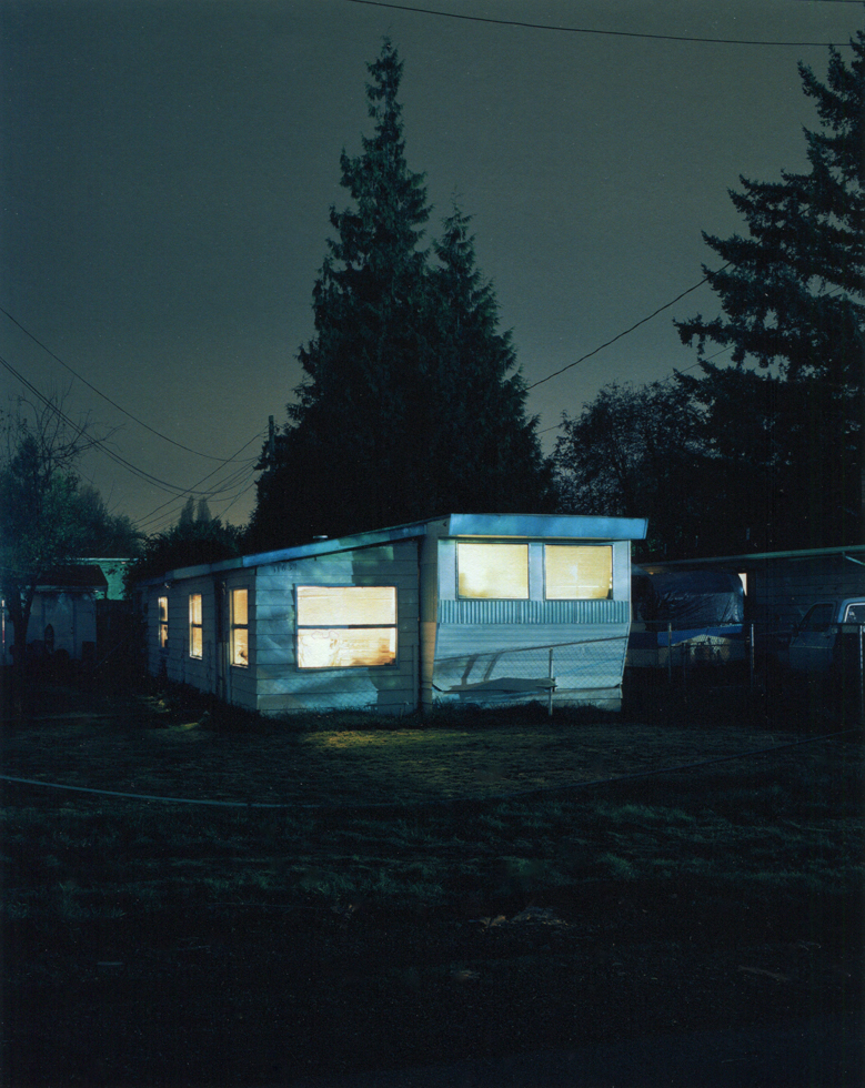 A trailer at night glows with interior light, its pale siding and windows stark against surrounding trees and deep shadows.