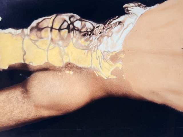 A colorful photograph of a nude man's buttocks and torso floating in the water with a reflection.