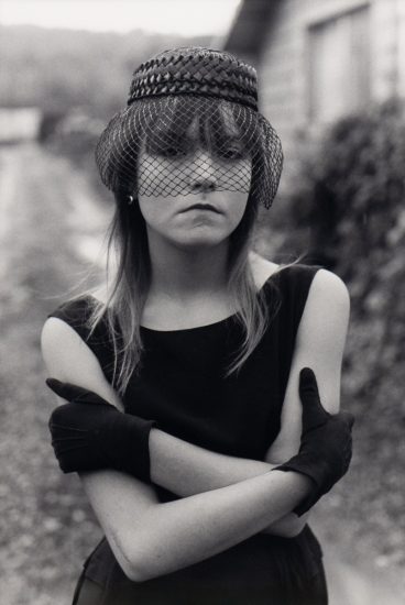 A woman in a sleeveless dress, netted hat, and gloves stands with crossed arms, gazing seriously in an outdoor setting.