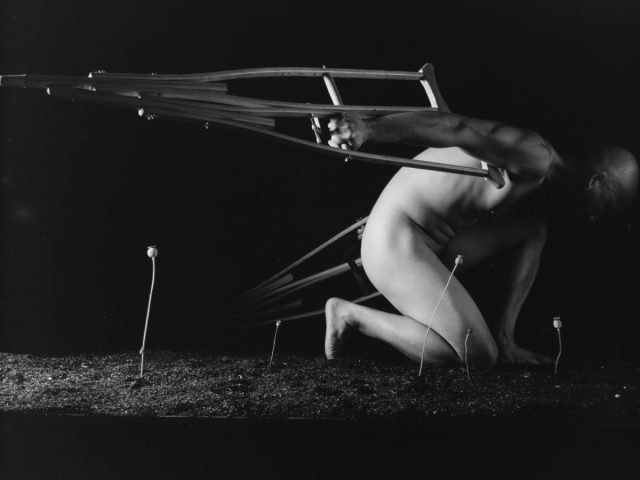 A bald, nude person crouches, with a pair of crutches attached to their back like wings, against a dark background with small plants growing.