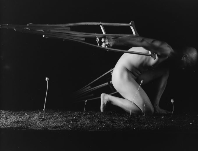 A bald, nude person crouches, with a pair of crutches attached to their back like wings, against a dark background with small plants growing.