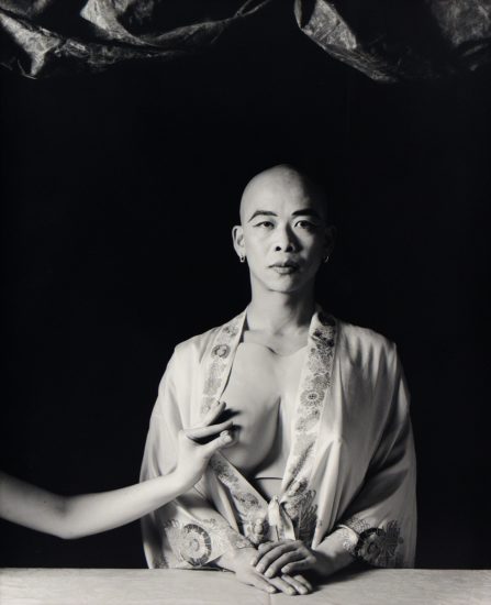 A black and white portrait of a bald person with their breast being pinched.