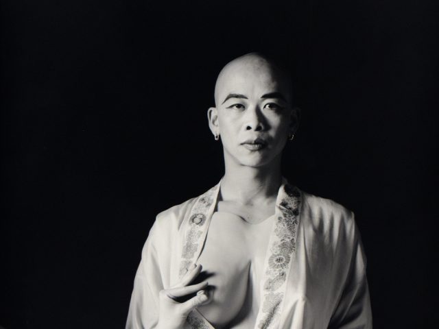 A black and white portrait of a bald person with their breast being pinched.