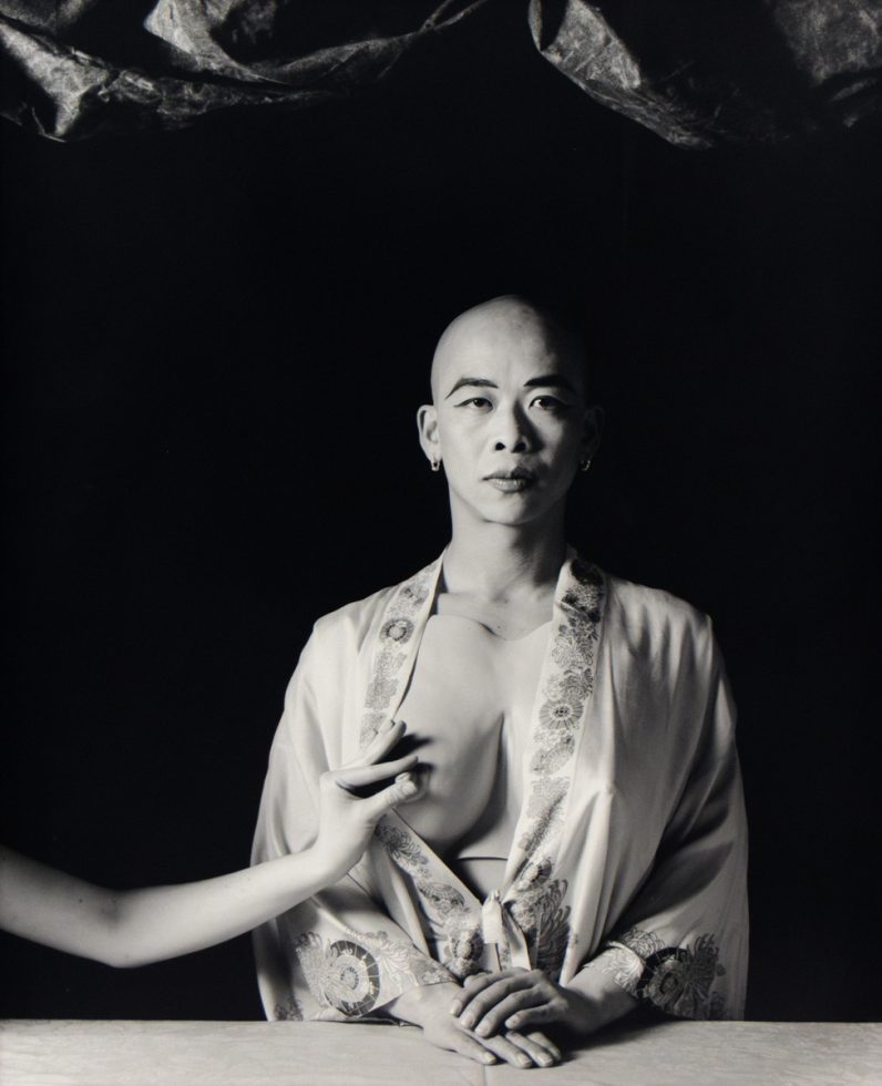 A black and white portrait of a bald person with their breast being pinched.