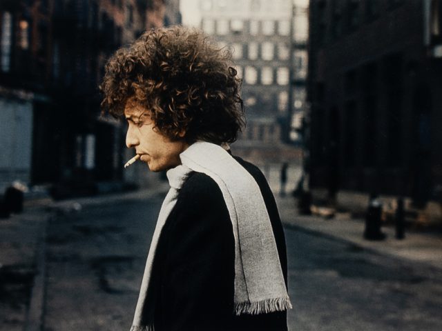 Man with curly hair and scarf smokes a cigarette while walking alone down a quiet, dimly lit city street.