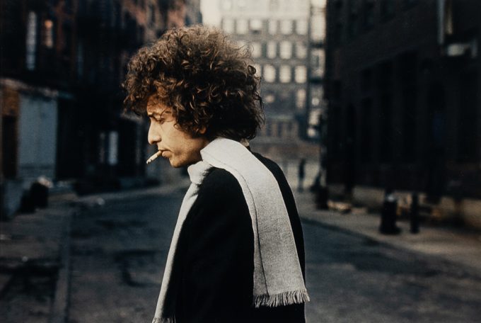 Man with curly hair and scarf smokes a cigarette while walking alone down a quiet, dimly lit city street.