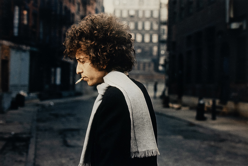 Man with curly hair and scarf smokes a cigarette while walking alone down a quiet, dimly lit city street.