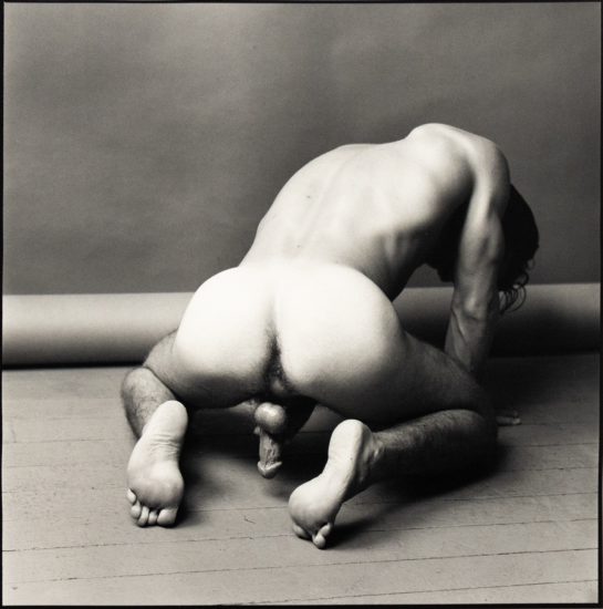 Black-and-white photograph of a nude figure kneeling forward on a studio floor, body bent and head turned down.