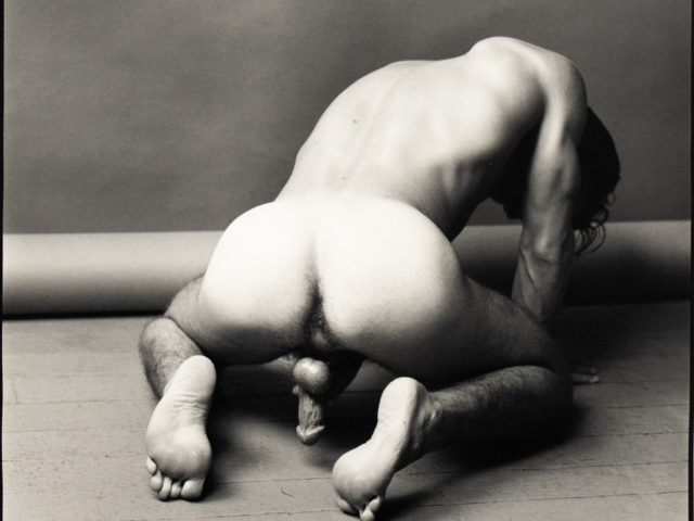 Black-and-white photograph of a nude figure kneeling forward on a studio floor, body bent and head turned down.
