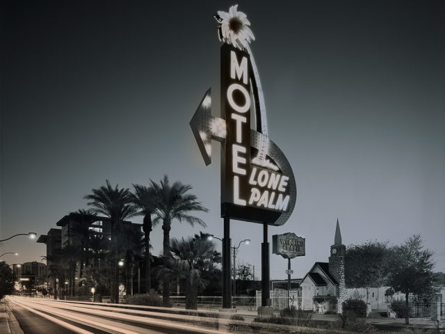 A huge motel sign with glowing neon letters on the side of a main road with blurry car lights.